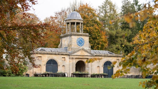 View of a large stone building with a huge clock on it. Autumnal trees in the background.